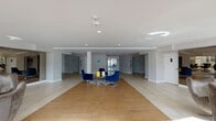 This single-floor space presents a well-appointed building lobby designed with a clean, modern aesthetic. The main entrance area features a combination of large-format porcelain tile and warm wood-tone flooring, creating a welcoming and visually dynamic foundation throughout the space. Recessed lighting illuminates the open layout, which is anchored by a central seating arrangement of bold navy blue swivel chairs gathered around a glass-topped coffee table atop a textured area rug.

Flanking the central lounge are additional seating vignettes furnished with sculptural egg-style chairs in taupe and gray, adding a contemporary design touch. A large bank of individual mailboxes lines one wall, serving as a functional focal point. Two elevators are accessible from the main lobby, providing vertical access to the broader building.

A glass-enclosed common room or amenity lounge connects to the lobby, visible through a mirrored sliding door. This bright secondary space benefits from expansive floor-to-ceiling windows that flood the interior with natural light and offer views of the surrounding outdoor greenery. The room is furnished with round tables and chairs, suggesting use as a social or meeting area. Together, these spaces combine practicality with a polished, residential-grade design sensibility.