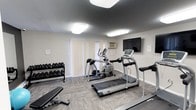 This fitness center is a compact yet fully functional workout space designed to support a variety of exercise routines. The room features durable, patterned carpet flooring that provides traction and comfort underfoot. Cardio equipment takes center stage, with a treadmill and an elliptical machine positioned prominently on the floor, catering to both walking and low-impact training needs. A weight bench and a well-stocked dumbbell rack line one side of the room, offering a solid range of free weights for strength training exercises. A wall-mounted flat-screen television is positioned at an ideal viewing angle, allowing users to stay entertained during their workouts. Natural light filters into the space through vertical blinds covering a large window, while flush-mounted ceiling lights ensure bright, even illumination throughout. A wall-mounted air conditioning unit helps maintain a comfortable temperature during intense sessions. The neutral, light-gray walls and clean layout give the space an organized and functional feel, making it easy to navigate and use efficiently. Overall, this fitness center provides a practical and accessible amenity for daily exercise.