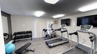 This fitness center is a compact yet fully functional workout space designed to support a variety of exercise routines. The room features durable, patterned carpet flooring that provides traction and comfort underfoot. Cardio equipment takes center stage, with a treadmill and an elliptical machine positioned prominently on the floor, catering to both walking and low-impact training needs. A weight bench and a well-stocked dumbbell rack line one side of the room, offering a solid range of free weights for strength training exercises. A wall-mounted flat-screen television is positioned at an ideal viewing angle, allowing users to stay entertained during their workouts. Natural light filters into the space through vertical blinds covering a large window, while flush-mounted ceiling lights ensure bright, even illumination throughout. A wall-mounted air conditioning unit helps maintain a comfortable temperature during intense sessions. The neutral, light-gray walls and clean layout give the space an organized and functional feel, making it easy to navigate and use efficiently. Overall, this fitness center provides a practical and accessible amenity for daily exercise.