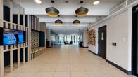 This thoughtfully designed single-floor amenity space combines modern aesthetics with practical functionality across several distinct areas. The expansive lobby entrance sets the tone immediately, featuring terrazzo tile flooring, exposed ceiling infrastructure, and a series of bold black-and-gold pendant lights that draw the eye down a generously proportioned corridor. A sculptural wood-slat bench with a colorful mosaic tile backing provides a striking focal point on one side, while an artistic wall installation of geometric wood pieces adds texture and visual interest on the opposite wall. Glass entry doors flood the entrance with natural light and offer views to the greenery outside. Moving deeper into the space, a stylish lounge area offers a curated residential feel, anchored by a floor-to-ceiling teal tile fireplace surround with built-in shelving, a wall-mounted television, and an eclectic mix of seating including velvet accent chairs in warm amber and navy tones. A private nook with tufted booth seating provides an intimate retreat within the lounge. The dining and social area features a large white communal table with modern seating and a bar-height counter with designer stools, all oriented toward expansive floor-to-ceiling glass doors that open to an outdoor terrace, allowing abundant natural light and seamless indoor-outdoor connection. A manager's office with glass partitioning completes the space near the main entry vestibule.
