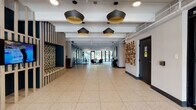 This thoughtfully designed single-floor amenity space combines modern aesthetics with practical functionality across several distinct areas. The expansive lobby entrance sets the tone immediately, featuring terrazzo tile flooring, exposed ceiling infrastructure, and a series of bold black-and-gold pendant lights that draw the eye down a generously proportioned corridor. A sculptural wood-slat bench with a colorful mosaic tile backing provides a striking focal point on one side, while an artistic wall installation of geometric wood pieces adds texture and visual interest on the opposite wall. Glass entry doors flood the entrance with natural light and offer views to the greenery outside. Moving deeper into the space, a stylish lounge area offers a curated residential feel, anchored by a floor-to-ceiling teal tile fireplace surround with built-in shelving, a wall-mounted television, and an eclectic mix of seating including velvet accent chairs in warm amber and navy tones. A private nook with tufted booth seating provides an intimate retreat within the lounge. The dining and social area features a large white communal table with modern seating and a bar-height counter with designer stools, all oriented toward expansive floor-to-ceiling glass doors that open to an outdoor terrace, allowing abundant natural light and seamless indoor-outdoor connection. A manager's office with glass partitioning completes the space near the main entry vestibule.