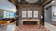 Located at 471 26th Street in Oakland, CA, this single-floor communal space masterfully blends industrial-rustic and contemporary design. The hallway entry makes an immediate impression with its reclaimed wood accent walls, an intricate laser-cut ceiling medallion, a tufted leather bench, and a trio of framed artwork — setting an artistic tone throughout.

The open living and kitchen area extends generously, offering several distinct zones for gathering and relaxation. Polished concrete floors run throughout, providing a sleek, cohesive foundation. Exposed dark steel beams frame the space, while reclaimed wood walls add warmth and texture. Pendant lighting in brass and globe styles casts a welcoming glow over a live-edge communal table surrounded by eclectic seating.

The lounge zone features deep channel-tufted sofas flanking a gold coffee table, with teal built-in shelving and a mounted television as the focal point. Floor-to-ceiling glass sliding doors flood the area with natural light and open directly onto a landscaped courtyard. A compact kitchen features bold teal cabinetry, white quartz countertops, a subway tile backsplash, open shelving, and stainless appliances — ideal for community gatherings. The overall ambiance is curated, vibrant, and welcoming.