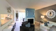 This single-floor residence offers a comfortable and stylishly appointed living space with a warm, inviting ambiance. The living room serves as the heart of the home, centered around a classic fireplace framed by a striking blue accent wall adorned with decorative wall art and candle sconces — creating a bold focal point against the neutral gray and beige tones throughout. Plush wall-to-wall carpeting adds warmth underfoot, while the open layout allows the living area to flow seamlessly into the dining space. Large sliding glass doors flood the interior with generous natural light and provide a direct view to an outdoor area, enhancing the sense of openness. The dining area accommodates a full-sized table and chairs, making it ideal for everyday meals or entertaining. A tall potted plant near the entrance adds a touch of greenery and life to the space. The interior is finished with textured ceilings and neutral-toned walls that complement the contemporary furnishings. Overall, this residence balances modern design elements with cozy, livable touches, making it a practical and aesthetically pleasing space.