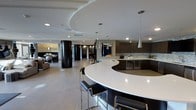 This single-floor space showcases a striking contemporary design defined by clean lines, high-end finishes, and thoughtful architectural elements. The centerpiece of the space is a sweeping, curved white kitchen island that commands attention, offering generous seating with modern black-and-chrome barstools. Overhead, sculptural tiered ceiling details add dimension and visual interest, complemented by sleek pendant lights in a brushed metallic finish that illuminate the workspace below.

The kitchen features dark espresso cabinetry paired with a stone mosaic backsplash, creating a sophisticated contrast against the bright white countertops. A built-in wine or beverage cooler is integrated seamlessly into the cabinetry, adding both function and style.

Dramatic cylindrical black columns punctuate the open-concept layout, reinforcing the bold design aesthetic while defining distinct zones within the space. The light hardwood flooring flows continuously throughout, enhancing the sense of openness. In the background, a lounge or living area is visible, featuring a fireplace and textured accent wall, contributing to the space's upscale, cohesive ambiance. Recessed lighting throughout ensures even illumination across the entire floor plan.