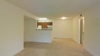 This single-floor residential unit offers a practical and functional layout designed for comfortable everyday living. The spacious living area is finished with neutral-toned carpet flooring and clean, light-colored walls that create a bright and airy atmosphere. Ceiling-mounted light fixtures provide consistent overhead illumination throughout the space.

The kitchen is positioned adjacent to the living area and is defined by a half-wall pass-through that doubles as a breakfast bar, creating a sense of openness while maintaining a distinct cooking zone. The kitchen features rich, dark wood cabinetry that contrasts with the lighter surroundings, along with a dark laminate countertop. A white refrigerator and under-cabinet lighting add to the kitchen's functionality. A skylight above the kitchen brings in additional natural light, further brightening the cooking space.

The overall design of the unit is straightforward and efficient, with a neutral color palette throughout that lends itself well to a variety of furnishing styles. The open-plan connection between the kitchen and living area enhances the sense of space, making the unit feel welcoming and versatile.