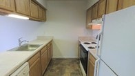 This two-floor space includes a galley-style kitchen that exemplifies a straightforward, functional design. The kitchen is laid out along two parallel walls, maximizing efficiency within a compact footprint. Warm-toned wooden cabinetry lines both the upper and lower sections of the walls, providing ample storage for pantry items and cookware. The countertops are finished in a neutral laminate, offering practical workspace on both sides of the kitchen. A stainless steel sink is positioned along the left counter run, while the right side accommodates a white coil-burner electric range with an overhead exhaust fan for ventilation. A white refrigerator anchors the far right end of the kitchen, completing the suite of standard appliances. The flooring features a durable, stone-patterned vinyl tile in earthy tones that complements the cabinetry. The walls are finished in a clean, neutral white, contributing to a bright and tidy atmosphere. The galley configuration ensures that all key work zones — refrigeration, preparation, cooking, and cleaning — are within close proximity, making the kitchen highly practical for daily use across both floors of the residence.