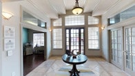 This lobby makes a striking first impression with its generous ceiling height, accentuated by tall arched transom windows flanking a set of dark-framed French entry doors. The abundance of glazing allows natural light to pour into the space, illuminating the warm neutral walls and decorative crown molding that lines the upper perimeter.

The flooring is a standout feature, consisting of large-format light tile arranged in a classic pattern with an inlaid geometric design at the center, adding a refined decorative touch. A dark wood round pedestal table anchors the center of the room, styled with tasteful décor accents that reinforce the polished aesthetic.

Wall sconces on either side of the entry doors provide a warm, ambient glow that complements the central pendant light fixture above, which features a classic bronze finish and frosted glass shade. An adjacent room, visible through an open doorway to the left, hints at additional functional office or administrative space. Overall, the lobby conveys a sense of elegance and openness, serving as a sophisticated transitional space.
