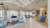 This single-story clubhouse showcases a coastal aesthetic defined by soaring vaulted ceilings with exposed white-painted beams and an abundance of natural light streaming through expansive floor-to-ceiling windows. The open-plan main lounge is anchored by light wood-tone flooring in a herringbone pattern and furnished with a sophisticated palette of navy, aqua, and neutral tones, complemented by ocean-inspired area rugs and pendant barn lights overhead.

The lounge flows seamlessly into a dining and gathering area, where a stone-clad accent wall hosts a sleek linear fireplace and wall-mounted television. Adjacent to this is a fully appointed kitchen featuring white cabinetry, quartz countertops, sage green tile backsplash, geometric tile flooring, a large center island with bar seating, and a built-in coffee station.

A dedicated workspace area, enclosed by glass-panel sliding doors, provides a quiet retreat with workstations and ample natural light. A glass-walled fitness center is visible from the main lounge, offering transparency while maintaining functionality. Throughout, the design maintains a cohesive, resort-style ambiance with views of palm trees and an outdoor patio, reinforcing the space's relaxed, coastal character.