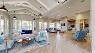 This single-story clubhouse showcases a coastal aesthetic defined by soaring vaulted ceilings with exposed white-painted beams and an abundance of natural light streaming through expansive floor-to-ceiling windows. The open-plan main lounge is anchored by light wood-tone flooring in a herringbone pattern and furnished with a sophisticated palette of navy, aqua, and neutral tones, complemented by ocean-inspired area rugs and pendant barn lights overhead.

The lounge flows seamlessly into a dining and gathering area, where a stone-clad accent wall hosts a sleek linear fireplace and wall-mounted television. Adjacent to this is a fully appointed kitchen featuring white cabinetry, quartz countertops, sage green tile backsplash, geometric tile flooring, a large center island with bar seating, and a built-in coffee station.

A dedicated workspace area, enclosed by glass-panel sliding doors, provides a quiet retreat with workstations and ample natural light. A glass-walled fitness center is visible from the main lounge, offering transparency while maintaining functionality. Throughout, the design maintains a cohesive, resort-style ambiance with views of palm trees and an outdoor patio, reinforcing the space's relaxed, coastal character.