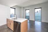 Located at 219 E Grand Ave in the heart of Des Moines, Iowa, this efficiently designed unit offers a clean, modern aesthetic throughout its single-floor layout. Polished concrete flooring runs through the main living area, lending an industrial-contemporary character that is both durable and visually striking.

The open living area is defined by floor-to-ceiling windows that flood the space with natural light and frame views of the surrounding urban landscape. Crisp white walls enhance the sense of openness and complement the minimalist design.

The kitchen is thoughtfully appointed with light maple cabinetry, white countertops, a central island with an integrated sink, and a full suite of stainless steel appliances including an electric range, microwave, refrigerator, and dishwasher. Recessed lighting provides focused illumination over the work and dining areas.

The bathroom features a combined tub and shower with tiled surround, a vanity with sink, and grab bar accessibility features, all finished in a clean, neutral palette. A dedicated closet provides convenient in-unit storage.

This unit's straightforward layout, modern finishes, and well-placed windows make it a practical and stylish option in downtown Des Moines.