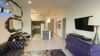 This single-floor space showcases an open-concept design that seamlessly connects the living, dining, and kitchen areas into one cohesive environment. The kitchen is equipped with dark cabinetry, stainless steel appliances, and a central island with seating, providing both functionality and a focal point for the space. Track lighting and recessed ceiling lights ensure the kitchen and adjoining areas are well-illuminated. The living area is defined by light-toned flooring that runs throughout, contributing to a clean and airy atmosphere. A wall-mounted flat-screen television anchors one side of the living space, accompanied by a striking purple dresser that adds a bold, eclectic character to the room. Decorative wall art featuring abstract patterns enhances the visual interest of the space. A small dining nook with a round table and chairs is thoughtfully integrated near the kitchen, offering a cozy spot for casual meals. A pendant light fixture above the kitchen island adds an elegant detail. The overall ambiance balances contemporary design with vibrant accent pieces, creating a space that feels both functional and personality-driven.