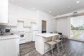This single-floor unit in Clarksville, TN offers a modern, minimalist aesthetic throughout its well-organized layout. The kitchen serves as a functional hub, featuring white cabinetry extending to the ceiling, a central island with a white countertop, and white appliances including a range, refrigerator, dishwasher, and microwave. Wood-look flooring runs through the kitchen and connects seamlessly to the living area, which includes a large floor-to-ceiling window fitted with horizontal blinds, providing views of the surrounding area and filling the space with natural light. A wall-mounted television adds a contemporary touch to the living space. The unit includes two bathrooms, each finished with wood-look flooring, white vanities, and clean neutral walls. One bathroom features a shower-tub combination with a framed mirror, while the other provides a more compact configuration. A carpeted area with a patterned tile-style carpet offers a distinct zone within the unit, complemented by a well-organized open closet system with shelving and hanging rods. An additional room with carpeting, a desk, and a window functions well as an office or study. The overall ambiance is bright, clean, and functional.