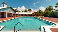 Located at 98 Paddock Club Drive in Panama City Beach, Florida, this outdoor amenity space offers a resort-style experience within a lush, tree-lined setting. The centerpiece of the facility is a generously sized swimming pool with crystal-clear turquoise water, accessible via wide entry steps and featuring clearly marked depth indicators — making it suitable for a range of swimmers. The pool is surrounded by an expansive brick-paved sun deck, providing ample room for lounge chairs and outdoor furnishings ideal for relaxing under the Florida sun. A covered pavilion with a distinctive metal roof and structural columns anchors one side of the deck, offering shaded seating and shelter from the elements. Adjacent to the pavilion, a pergola-style structure extends the covered outdoor living area, adding architectural character to the space. Bright yellow umbrellas are positioned throughout the deck, adding a pop of color while providing additional shade. The facility is bordered by a privacy fence and rich tropical greenery, including palms and mature trees that frame the space and contribute to its secluded, resort-like atmosphere. A single-story clubhouse building with classic coastal architectural detailing completes the complex, tying together the functional and aesthetic elements of this well-appointed outdoor amenity area.