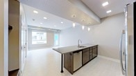 This thoughtfully designed single-floor space combines contemporary aesthetics with functional living. The open-concept layout seamlessly connects the kitchen and living areas, creating an inviting and airy atmosphere. A standout feature is the kitchen island, finished with a sleek dark countertop and equipped with a stainless steel sink and dishwasher. Dark cabinetry adds a sophisticated contrast to the otherwise light and neutral palette throughout the space. Above the island, an elegant tray ceiling section is adorned with a bold geometric patterned inset, adding a striking visual focal point. Crystal pendant lights hang gracefully from this section, lending warmth and a refined touch to the kitchen area. Recessed lighting is distributed throughout, ensuring consistent, well-balanced illumination across the space. The living area benefits from abundant natural light, courtesy of large windows that frame views of the surrounding exterior. Light-toned flooring flows continuously through both areas, reinforcing the sense of openness and cohesion. Clean white walls and minimalist finishes contribute to the space's modern, understated character, making it a versatile and welcoming environment.