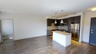 This single-floor space showcases a contemporary open-concept design that seamlessly integrates the kitchen and living areas into one cohesive environment. The kitchen is a standout feature, equipped with dark espresso cabinetry that contrasts elegantly against light stone countertops. A central island with a crisp white base provides additional prep space and serves as a natural gathering point. Stainless steel appliances, including a refrigerator, range, and microwave, add a polished, modern touch. Two brushed nickel pendant lights hang above the island, complementing a track lighting system that runs along the ceiling to illuminate the cooking space effectively. A tile backsplash adds texture and visual interest behind the counters. The open living area is finished with wide-plank hardwood-style flooring in a warm gray-brown tone, lending warmth and continuity throughout the space. Neutral white walls keep the interior bright and versatile, offering a clean backdrop adaptable to various décor styles. The overall ambiance is modern, clean, and functional, making it well-suited for everyday living.