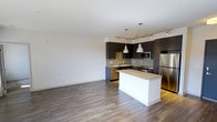 This single-floor space showcases a contemporary open-concept design that seamlessly integrates the kitchen and living areas into one cohesive environment. The kitchen is a standout feature, equipped with dark espresso cabinetry that contrasts elegantly against light stone countertops. A central island with a crisp white base provides additional prep space and serves as a natural gathering point. Stainless steel appliances, including a refrigerator, range, and microwave, add a polished, modern touch. Two brushed nickel pendant lights hang above the island, complementing a track lighting system that runs along the ceiling to illuminate the cooking space effectively. A tile backsplash adds texture and visual interest behind the counters. The open living area is finished with wide-plank hardwood-style flooring in a warm gray-brown tone, lending warmth and continuity throughout the space. Neutral white walls keep the interior bright and versatile, offering a clean backdrop adaptable to various décor styles. The overall ambiance is modern, clean, and functional, making it well-suited for everyday living.