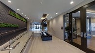 This single-floor lobby presents a refined and contemporary design, combining clean lines with upscale finishes to create an inviting entrance experience. The space is defined by light hardwood-style flooring that runs the full length of the corridor, complemented by smooth, neutral-toned walls and a bright white ceiling with recessed lighting throughout.

A standout centerpiece is the dark, marble-finished water feature positioned along the central walkway, providing both visual interest and a calming ambiance. Above it, elegant geometric pendant lights hang from the ceiling, adding a decorative focal point that enhances the modern aesthetic.

Glass railings line both sides of the walkway, contributing to the open and airy feel of the space. To one side, a living green wall installation introduces a natural element, adding texture and vibrancy to the otherwise neutral palette. On the opposite side, floor-to-ceiling glass doors lead to an adjacent room, where lush indoor plants and additional seating areas are visible.

At the far end, double glass entry doors and a staircase draw the eye forward, reinforcing the sense of depth and connectivity within the space. Overall, the lobby balances sophistication with functionality.