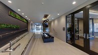 This single-floor lobby presents a refined and contemporary design, combining clean lines with upscale finishes to create an inviting entrance experience. The space is defined by light hardwood-style flooring that runs the full length of the corridor, complemented by smooth, neutral-toned walls and a bright white ceiling with recessed lighting throughout.

A standout centerpiece is the dark, marble-finished water feature positioned along the central walkway, providing both visual interest and a calming ambiance. Above it, elegant geometric pendant lights hang from the ceiling, adding a decorative focal point that enhances the modern aesthetic.

Glass railings line both sides of the walkway, contributing to the open and airy feel of the space. To one side, a living green wall installation introduces a natural element, adding texture and vibrancy to the otherwise neutral palette. On the opposite side, floor-to-ceiling glass doors lead to an adjacent room, where lush indoor plants and additional seating areas are visible.

At the far end, double glass entry doors and a staircase draw the eye forward, reinforcing the sense of depth and connectivity within the space. Overall, the lobby balances sophistication with functionality.