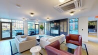 This thoughtfully designed single-floor amenity space offers a sophisticated blend of comfort and contemporary style across two distinct zones. The main lounge area is anchored by a striking dark-wood built-in entertainment wall, complete with a sleek linear fireplace and a mounted flat-screen TV flanked by glass-front cabinetry. Plush seating arrangements — including neutral sofas, leather accent chairs, and modern side tables — are oriented around this focal point, creating an inviting atmosphere for relaxation or socializing. Light hardwood flooring, recessed lighting, and flush-mount globe fixtures contribute to the warm, refined ambiance throughout.

Floor-to-ceiling windows and glass doors bathe the lounge in abundant natural light while offering direct views and access to an outdoor terrace. Lush potted plants and greenery are thoughtfully placed throughout, adding a natural, organic quality to the interior.

A second zone features a vibrant dining and lounge area distinguished by bold mustard-yellow tufted banquette seating, dark marble-topped tables, and mixed wood and upholstered chairs. The ceiling in this area is dramatically adorned with cascading greenery and hanging globe lights set against dark-painted structural beams, creating an immersive, garden-inspired atmosphere. Together, the two areas form a cohesive and dynamic communal space designed for both comfort and community engagement.