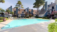 This thoughtfully designed single-floor residence is part of a well-established multi-unit community characterized by classic pitched rooflines, blue and neutral exterior tones, and lush landscaping anchored by towering pine trees. The community's centerpiece is a generously sized outdoor swimming pool, lined with lounge chairs and a spacious concrete deck — ideal for relaxation and recreation.

Inside, the interior reflects a curated, contemporary aesthetic. The dining area serves as a standout space, anchored by a striking round marble-topped table in deep green tones, paired with rattan-style chairs and set atop a woven area rug. Dark hardwood flooring grounds the room, while recessed lighting illuminates the clean white walls, which are adorned with large-scale framed artwork that adds an artistic, gallery-like quality to the space.

A distinctive sliding barn door with a matte black rail system adds an industrial yet refined touch, offering a stylish transition between rooms. An adjacent doorway fitted with vintage-style paneled glass doors connects to a neighboring room displaying framed wall art, hinting at a creative and personalized living environment throughout.

The overall ambiance is calm and sophisticated, blending warm textures, curated décor, and architectural details to create a space that feels both livable and visually engaging.