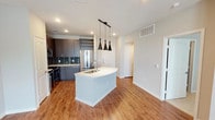 This well-appointed single-floor residence showcases a thoughtfully designed open-concept layout that seamlessly connects the kitchen and living areas. The kitchen is a standout feature, boasting dark wood cabinetry with sleek hardware, a teal glass tile backsplash, and a full suite of stainless steel appliances including a French door refrigerator, electric range, and over-the-range microwave. A curved center island with a white countertop and undermount sink serves as both a functional workspace and a focal point, illuminated by three striking black pendant lights. A built-in wine rack above the refrigerator adds a sophisticated touch. Warm wood-look flooring flows throughout the main living spaces, including the living room, which opens to a private balcony through glass French doors and is finished with a ceiling fan. The bedrooms feature plush carpeting, ceiling fans, and windows with blinds that allow for natural light. The bathroom is elegantly finished with large-format tile flooring, a granite-topped double vanity with dark wood cabinetry, and a glass-enclosed shower with dark tile surround. Recessed lighting and neutral wall tones throughout the space create a clean, contemporary ambiance.