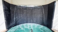 This single-level space centers around a standout hydrotherapy feature: a generously sized circular hot tub with active jets producing visibly bubbling, turquoise-tinted water. The most commanding element is the curved, dark textured wall that wraps around the tub in a near-semicircular formation, from which cascading water streams flow continuously downward, creating a full waterfall curtain effect. The wall's dark, rough-hewn surface contrasts dramatically with the luminous water, enhancing the immersive atmosphere. Above the waterfall wall, an opening allows natural light to enter the space, with glimpses of tropical or bamboo-like foliage visible beyond, connecting the interior to the outdoors. The overall design evokes a grotto-like sensory environment, blending natural stone-inspired textures with contemporary spa engineering. The enclosure creates a sense of privacy and seclusion, making the space feel like a dedicated wellness retreat. The continuous sound and motion of the cascading water, combined with the jetted pool below, contribute to a deeply relaxing and visually captivating ambiance suited for therapeutic or leisure use.