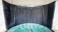 This single-level space centers around a standout hydrotherapy feature: a generously sized circular hot tub with active jets producing visibly bubbling, turquoise-tinted water. The most commanding element is the curved, dark textured wall that wraps around the tub in a near-semicircular formation, from which cascading water streams flow continuously downward, creating a full waterfall curtain effect. The wall's dark, rough-hewn surface contrasts dramatically with the luminous water, enhancing the immersive atmosphere. Above the waterfall wall, an opening allows natural light to enter the space, with glimpses of tropical or bamboo-like foliage visible beyond, connecting the interior to the outdoors. The overall design evokes a grotto-like sensory environment, blending natural stone-inspired textures with contemporary spa engineering. The enclosure creates a sense of privacy and seclusion, making the space feel like a dedicated wellness retreat. The continuous sound and motion of the cascading water, combined with the jetted pool below, contribute to a deeply relaxing and visually captivating ambiance suited for therapeutic or leisure use.