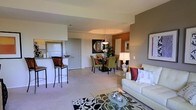 This single-floor residence showcases an open-concept design that integrates the living, dining, and kitchen areas into one cohesive, flowing space. The living area is anchored by a comfortable white sofa accented with burgundy and patterned throw pillows, complemented by a glass coffee table resting atop a bold black-and-white floral area rug. An accent wall painted in a warm terracotta tone adds depth and visual interest to the space.

The dining area features a dark wood table surrounded by green upholstered chairs, positioned beneath an elegant pendant light with a tray ceiling detail that draws the eye upward. A decorative mirror and framed artwork on the accent wall enhance the room's character.

The kitchen is partially open to the main living space via a raised breakfast bar with bar stool seating, offering a convenient casual dining option. Granite countertops and standard appliances are visible from the main area. Light tile flooring in the kitchen transitions smoothly to plush neutral carpeting throughout the living and dining zones. The overall ambiance is warm, stylish, and well-appointed.