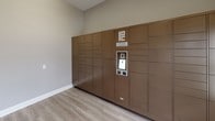 This Package Concierge room is a thoughtfully designed, single-floor space dedicated to secure and convenient parcel retrieval. The room is finished with light wood-style luxury vinyl flooring and neutral warm-gray walls, creating a clean and welcoming environment. Dominating one full wall is a large bank of bronze-toned automated Package Concierge lockers, offering a wide range of compartment sizes to accommodate deliveries of varying dimensions. The locker system features a central digital interface, allowing residents to retrieve packages with ease at any time of day. A stylish hexagonal accent table with a white marble top and gold base is positioned in the corner of the room, displaying informational signage to guide users. Natural light filters into the space through a double-hung window, and a glass-paneled entry door provides secure yet accessible entry to the room. A fire safety panel is also mounted on the wall near the entry. The overall design of the space balances functionality with a polished, modern aesthetic, ensuring that package management is both efficient and seamlessly integrated into the residential experience.