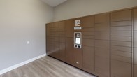 This Package Concierge room is a thoughtfully designed, single-floor space dedicated to secure and convenient parcel retrieval. The room is finished with light wood-style luxury vinyl flooring and neutral warm-gray walls, creating a clean and welcoming environment. Dominating one full wall is a large bank of bronze-toned automated Package Concierge lockers, offering a wide range of compartment sizes to accommodate deliveries of varying dimensions. The locker system features a central digital interface, allowing residents to retrieve packages with ease at any time of day. A stylish hexagonal accent table with a white marble top and gold base is positioned in the corner of the room, displaying informational signage to guide users. Natural light filters into the space through a double-hung window, and a glass-paneled entry door provides secure yet accessible entry to the room. A fire safety panel is also mounted on the wall near the entry. The overall design of the space balances functionality with a polished, modern aesthetic, ensuring that package management is both efficient and seamlessly integrated into the residential experience.