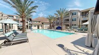Located at 21595 Box Springs Rd in Moreno Valley, CA, this outdoor amenity space offers a resort-inspired retreat set against the striking backdrop of Box Springs Mountain. The centerpiece is a generously sized swimming pool with crystal-clear water, surrounded by a broad, well-maintained concrete pool deck ideal for lounging and socializing under the Southern California sun.

Tall, mature palm trees are strategically placed throughout the area, providing natural shade and reinforcing the tropical, resort-like atmosphere. Multiple chaise lounges and poolside seating are arranged along the deck, offering ample space for relaxation. Patio umbrellas provide additional shade at key gathering spots.

A standout feature of the space is the dedicated cabana area, equipped with draped canopy structures offering privacy and shade. Each cabana is furnished with plush, teal-cushioned seating and includes a mounted flat-screen television, combining comfort with modern amenities. The open-air cabanas are further enhanced by ceiling fans and elegant draping curtains that can be drawn for a more intimate setting.

The surrounding multi-story residential buildings feature Spanish-influenced architecture with terracotta tile rooflines, stucco exteriors, and decorative ironwork railings. Lush, drought-tolerant plantings line the perimeter, tying together the landscaping in a cohesive, desert-friendly design that complements the natural mountain scenery beyond.