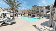 Located at 21595 Box Springs Rd in Moreno Valley, CA, this outdoor amenity space offers a resort-inspired retreat set against the striking backdrop of Box Springs Mountain. The centerpiece is a generously sized swimming pool with crystal-clear water, surrounded by a broad, well-maintained concrete pool deck ideal for lounging and socializing under the Southern California sun.

Tall, mature palm trees are strategically placed throughout the area, providing natural shade and reinforcing the tropical, resort-like atmosphere. Multiple chaise lounges and poolside seating are arranged along the deck, offering ample space for relaxation. Patio umbrellas provide additional shade at key gathering spots.

A standout feature of the space is the dedicated cabana area, equipped with draped canopy structures offering privacy and shade. Each cabana is furnished with plush, teal-cushioned seating and includes a mounted flat-screen television, combining comfort with modern amenities. The open-air cabanas are further enhanced by ceiling fans and elegant draping curtains that can be drawn for a more intimate setting.

The surrounding multi-story residential buildings feature Spanish-influenced architecture with terracotta tile rooflines, stucco exteriors, and decorative ironwork railings. Lush, drought-tolerant plantings line the perimeter, tying together the landscaping in a cohesive, desert-friendly design that complements the natural mountain scenery beyond.