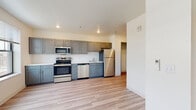 This single-floor unit offers a clean, contemporary design with a thoughtful layout and quality finishes throughout. The open-concept living and kitchen area is anchored by light-toned luxury vinyl plank flooring that extends seamlessly across the space, creating a bright and cohesive feel. Recessed lighting illuminates the interior, complemented by natural light streaming in through multiple windows that frame views of the surrounding urban streetscape.

The kitchen is a standout feature, showcasing gray shaker-style cabinetry on both upper and lower sections, paired with light marble-look countertops and a white subway tile backsplash. A full suite of stainless steel appliances includes a refrigerator, range with oven, over-the-range microwave, and dishwasher, making the kitchen as functional as it is stylish.

The bedroom offers plush wall-to-wall carpeting and is bathed in natural light from dual windows, providing open views of the cityscape beyond. The bathroom is finished with a sleek, frameless sliding glass walk-in shower enclosure set against clean white tile, accented by brushed nickel hardware, and continues the light wood-look flooring found throughout the unit. Overall, the space presents a modern, move-in-ready environment with a neutral palette and urban character.