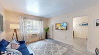 This single-floor unit offers a thoughtfully arranged layout that balances comfort and functionality. The living area serves as the heart of the space, featuring light wood-style flooring, neutral warm-toned walls, and a large window dressed with patterned curtains that allow generous natural light to fill the room. A wall-mounted flat-screen TV, a plush area rug, and a vibrant blue sofa anchor the space, creating an inviting atmosphere. A wall-unit air conditioner and a louvered storage unit contribute to the room's practical design.

The kitchen is efficiently designed, showcasing white cabinetry, dark granite-style countertops, and a stainless steel sink. An arched decorative mirror adds a charming architectural accent, while under-cabinet lighting enhances the workspace.

The bedroom is a restful retreat, highlighted by ombre blue curtains framing a natural-light window, elegant wall-mounted sconce lighting on either side of the bed, and coordinating blue-toned bedding. A louvered unit provides additional storage, and framed wall art adds a personalized touch.

Throughout the unit, the consistent use of blue accents, wood-style flooring, and soft neutral walls creates a cohesive and welcoming aesthetic suited for comfortable everyday living.