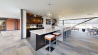 Located at 510 Taylor Place in Nashville, Tennessee, this single-floor community amenity space is designed to encourage socializing and relaxation in a polished, contemporary setting. The open layout flows seamlessly between distinct functional zones, unified by patterned, multi-toned carpet tiles that add texture and visual depth throughout.

The lounge area serves as the centerpiece, anchored by a generous white sectional sofa and complementary seating arrangements. Warm exposed brick accent walls provide an industrial contrast to the otherwise modern, white ceiling and recessed lighting. A wall-mounted flat-screen television enhances the entertainment options available in this area.

Adjacent to the lounge, a fully equipped kitchen features dark cabinetry, a stainless steel refrigerator, microwave, and two expansive islands — one with a white quartz surface and the other finished in a warm gray tone — each lined with stylish bar seating beneath globe pendant lights. A built-in television and stone surround add a refined focal point to the kitchen zone.

A dedicated game area includes an octagonal game table surrounded by contemporary chairs, positioned near the brick accent wall. Floor-to-ceiling windows line the perimeter, flooding the space with natural light and offering views of the surrounding pool and courtyard area, creating a bright and inviting atmosphere throughout.