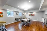 This well-appointed single-floor space offers a thoughtfully designed open-plan layout that seamlessly integrates multiple functional zones. The main area is anchored by warm hardwood flooring throughout, complemented by neutral gray-white walls and a combination of recessed lighting and a central flush-mount ceiling fixture that together create a bright, welcoming atmosphere.

The central gathering area features a large white rectangular table surrounded by modern chairs, suitable for meetings or group activities. Adjacent to the table, a dedicated lounge zone includes comfortable seating chairs arranged along the windowed wall, a wall-mounted flat-screen television, and a media console — offering a relaxed setting for informal use. Multiple windows along the perimeter flood the space with natural light and provide views of the surrounding outdoors.

The fully equipped kitchen is outfitted with crisp white shaker-style cabinetry, granite-look countertops, and a complete suite of stainless steel appliances including a refrigerator, electric range, microwave, and dishwasher. The kitchen opens directly to the main gathering area, making it highly functional for events or catered use.

Colorful artwork adorns the walls throughout, adding character and visual interest. A wall-mounted clock, an HVAC unit, and a water dispenser near the entry round out the practical amenities of this well-designed, multifunctional space.