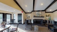 This single-floor space is designed as a versatile communal lounge, combining several distinct yet open seating areas under one roof. The architecture is defined by bold dark-stained exposed beams that contrast against textured white ceilings, lending a structured, elegant character throughout. A coffered ceiling section fitted with skylights and a ceiling fan floods the central lounge area with natural light, enhancing its welcoming ambiance.

The main seating zone features a large dark leather sectional sofa arranged around a wall-mounted television, creating a relaxed media lounge atmosphere. Adjacent to this is a more formal sitting area anchored by a black mosaic tile fireplace with a dark mantel shelf, accompanied by upholstered wingback chairs and a decorative accent table. Contemporary artwork adorns the walls, complementing the neutral beige tones and dark trim throughout.

A dining area near the rear of the space benefits from an abundance of natural light streaming through multiple sets of French doors that open to an outdoor deck with railing views beyond. The patterned carpet flows consistently throughout, unifying the varied zones. A classic wood-framed French interior door connects separate sections of the space, adding architectural charm and visual continuity.