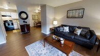 This single-floor Cumberland residence offers two bedrooms and two bathrooms across a generously sized floor plan. The living area serves as the heart of the home, featuring rich dark hardwood flooring that extends throughout the main living spaces. Neutral gray walls provide a versatile backdrop, complementing the warm wood tones and classic décor. A comfortable seating area anchors the living room, accented by a patterned area rug and a wood coffee table. Decorative touches, including a round accent mirror and a landscape art piece, add character to the space. A partial wall column subtly defines the transition between the living room and the adjacent dining area, which is visible in the background and accommodates a full dining set beneath warm overhead lighting. The open sightlines between rooms enhance the sense of spaciousness and connectivity throughout the home. The overall ambiance is warm and inviting, combining practical layout with tasteful interior choices, making this residence a comfortable and well-appointed living space suitable for a variety of lifestyles.