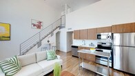 This contemporary two-story space is defined by its dramatic double-height ceilings and open-concept design, creating an airy, expansive atmosphere throughout. The main level integrates the living and kitchen areas in a cohesive, functional layout. The living area features light hardwood flooring, clean white walls, and a comfortable neutral-toned sofa accented with vibrant greenery, contributing to a fresh, modern aesthetic. A sleek stainless steel kitchen island anchors the cooking space, which is equipped with stainless steel appliances, including a built-in microwave, electric range, and dishwasher. Warm wood-toned cabinetry lines the kitchen walls, providing ample storage while adding natural warmth to the otherwise crisp, minimalist palette. A modern metal-railed staircase rises from the main floor to the upper level, its open design reinforcing the sense of vertical space and allowing light to flow freely between floors. The upper level is visible from below, overlooking the main living area through the open railing. Throughout the space, the interplay of warm wood tones, stainless steel finishes, and bright white surfaces creates a balanced, contemporary environment that feels both functional and inviting.