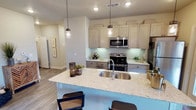 This single-floor residence showcases a thoughtfully designed open-concept kitchen that serves as the centerpiece of the living space. The kitchen is anchored by a large island topped with light-speckled granite countertops, which doubles as a casual dining area with modern bar-height seating. A deep double-basin stainless steel sink is integrated into the island, complemented by a sleek contemporary faucet.

The cabinetry features a warm greige finish with clean lines, extending to the upper walls and providing generous storage throughout. Stainless steel appliances, including a range with an overhead microwave, add a polished, modern touch. Under-cabinet lighting illuminates the countertop workspace, enhancing both functionality and ambiance.

Three pendant lights with Edison-style bulbs hang above the island, offering focused task lighting with a stylish aesthetic. Recessed ceiling lights provide additional illumination throughout. The flooring consists of wood-look planks in a neutral tone, contributing warmth and continuity across the open layout. A hallway visible beyond the kitchen suggests well-connected living spaces throughout the unit, reinforcing its practical and cohesive design.