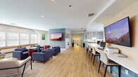Located at 100 Hunsberger Dr in Limerick, PA, this well-appointed single-floor community clubhouse offers a thoughtfully designed layout that balances social gathering, productivity, and relaxation. The open-plan interior flows seamlessly between distinct functional zones, unified by warm hardwood-style flooring, recessed lighting, and a bright, neutral color palette.

The main lounge area is anchored by a large wall-mounted flat-screen TV set against a textured accent wall, complemented by plush navy blue armchairs arranged around game-top side tables. A shuffleboard table and an abundance of natural light from multiple white-trimmed windows enhance the recreational atmosphere. Adjacent to the lounge, a long bar-height workstation with computer monitors and bar seating offers a practical co-working space beneath modern pendant lighting.

The fully equipped kitchen features soft blue shaker-style cabinetry, white quartz countertops, stainless steel appliances including a dishwasher and beverage refrigerator, and a tiled island. Colorful artwork adorns the walls, adding vibrancy to the space. A cozy windowed nook with a round dining table provides an intimate spot for small gatherings. Throughout, the design strikes a balance between contemporary style and functional community living.