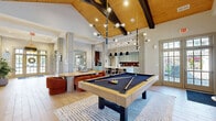 Located at 1100 Monacan Ln in Earlysville, VA, this expansive single-floor amenity space offers a thoughtfully designed collection of rooms suited for both relaxation and active living. The welcoming living room serves as the heart of the facility, showcasing soaring ceilings, a dramatic floor-to-ceiling dark brick fireplace with a mounted TV, a bold circular chandelier, and stylish mid-century modern furnishings atop a terrazzo-style floor. An adjacent dining room features a refined round table beneath an artisan chandelier, built-in cabinetry, floating shelves, and natural light from large windows. The kitchen and living room area is appointed with sage-toned shaker cabinetry, a rich burgundy tile backsplash, stainless steel appliances, a marble-topped island with pendant lighting, and light wood-look flooring that extends toward a glass-panel exterior door. The comprehensive exercise room is divided into distinct zones: a cardio area with treadmills, ellipticals, and stationary bikes beneath a warm wood coffered ceiling with ambient lighting; a strength training area with cable machines and free weights; and a dedicated spin and functional training studio with hardwood floors, cycling bikes, a full-length mirror, and ceiling fans. The bathroom is finished with herringbone wood-look tile wainscoting, slate-toned floor tiles, and ADA-compliant fixtures. Three hallways connect the rooms seamlessly throughout this well-appointed facility.