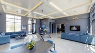 This refined single-floor space makes an immediate impression with its striking coffered ceiling, finished in crisp white and accented with warm recessed lighting that casts a soft, inviting glow across the entire interior. The open-plan layout is expansive and thoughtfully arranged, with distinct zones for lounging, dining, and socializing that flow seamlessly into one another. Cool grey walls provide a sophisticated backdrop that complements the light-toned herringbone-patterned flooring, lending the space a polished, contemporary character. The dining area is anchored by a sleek dark table with geometric X-shaped legs, surrounded by plush blue velvet chairs and adorned with elegant floral arrangements. A modern oval pendant light hangs above, adding a focal point to the dining zone. The lounge area features coordinating blue upholstered sofas arranged around a wall-mounted flat-screen display, creating a comfortable and stylish gathering space. Floor-to-ceiling black-framed glass doors and windows along one wall flood the interior with abundant natural light and offer views to an outdoor area beyond. Additional rooms visible through connecting doorways suggest further functional spaces, all maintaining the same cohesive, upscale aesthetic throughout.