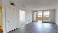 This single-floor residence showcases a thoughtfully designed open layout with a clean, contemporary aesthetic carried consistently throughout. The main living area features smooth wood-look flooring in a cool gray tone, crisp white walls, and a track light fixture overhead. A glass-paneled door leads directly to a private balcony, where views of palm trees and surrounding greenery are visible, bringing natural light deep into the living space.

The kitchen is well-equipped with warm wood-grain cabinetry spanning floor to ceiling, light quartz countertops, a white tile backsplash, and a full suite of stainless steel appliances including a gas range, built-in microwave, refrigerator, and dishwasher. The layout is efficient and functional with ample storage.

The bedrooms are carpeted for comfort and feature white walls with large windows that admit natural light. One bedroom includes a distinctive three-panel frosted glass sliding closet, adding a modern design touch. An additional room with wood-look flooring and a glass door offers flexibility as a secondary bedroom or office.

The bathroom is finished with light tile flooring, a wood-grain vanity with a white undermount sink, a large mirror, and a glass-enclosed tub and shower combination with tiled surround.
