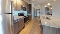 Located at 100 Whitworth Way in Williamsburg, Virginia, this well-appointed single-floor residence offers a thoughtfully designed layout with contemporary finishes throughout. The open-concept kitchen serves as a central highlight, showcasing rich dark-wood cabinetry, granite countertops, and a large center island with pendant lighting overhead. Stainless steel appliances, including a refrigerator, electric range, microwave, and dishwasher, complete the fully equipped kitchen. Light hardwood-style flooring runs through the main living and kitchen areas, contributing to a bright, cohesive aesthetic. The spacious living area features a ceiling fan and large windows that invite natural light and outdoor views. Multiple bedrooms are finished with plush carpeting, ceiling fans, and generous walk-in closets, providing ample storage. One bedroom benefits from multiple windows on two walls, filling the room with abundant natural light. The bathrooms are consistently finished with granite-topped dark wood vanities, undermount sinks, brushed nickel fixtures, and tub-shower combinations with tile surrounds. Hardwood-style flooring continues into the bathrooms, maintaining a uniform and modern look throughout. This residence combines functional design with quality finishes, creating a comfortable and stylish living environment.