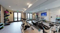 This purpose-built cycling studio is designed to deliver an immersive and high-energy workout experience. The room is outfitted with a row of professional-grade spinning bikes arranged to face a full-length wall mirror, allowing riders to monitor their form throughout each session. The mirrored wall also visually expands the space, enhancing the open and energetic atmosphere of the studio. A wall-mounted flat-screen display serves as a focal point for guided workouts or virtual cycling classes, adding a modern, tech-forward dimension to the fitness experience. The studio is finished with light hardwood-style flooring that complements the clean, contemporary aesthetic throughout. Recessed lighting in the white ceiling provides bright, even illumination across the room. A textured accent wall adds visual interest and depth to the space. Large glass-panel double doors open directly to an outdoor area, inviting natural light into the studio and offering a refreshing sense of openness. The doors also provide convenient ventilation during intense training sessions. An accessory rack mounted near the mirror offers organized storage for workout gear. Overall, the studio combines functionality and modern design to create a focused, motivating environment for cycling enthusiasts.