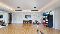 This fitness studio offers a thoughtfully designed workout space spread across a single floor. The room is finished with light wood-tone flooring that creates a clean, energetic atmosphere suited to active use. A tray ceiling with recessed cove lighting and suspended linear pendant fixtures adds architectural depth and provides well-distributed illumination throughout the space.

One wall is clad in a textured, whitewashed wood-style wallcovering, lending a modern yet warm aesthetic. A wall-mounted flat-screen television is centered on this feature wall, accompanied by a navy blue storage unit with woven basket organizers—ideal for storing small fitness accessories. A large wall mirror on an adjacent side enhances the sense of space and supports form-focused training.

Floor-to-ceiling windows with sheer white curtains line two sides of the room, inviting generous natural light and offering views of the surrounding greenery and outdoor environment. The windows also allow direct access via glass doors, enhancing ventilation and connectivity to the outdoors.

Equipment includes multiple spin bikes arranged for group cycling sessions, a dumbbell rack, medicine ball storage tower with an assortment of weighted balls, and additional free weights. The open floor plan allows for flexible use, accommodating both equipment-based and open-floor exercise activities.