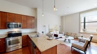 This single-story unit in New Orleans, LA, offers a thoughtfully designed open-concept layout that maximizes its efficient footprint. Soaring ceilings throughout the living and kitchen areas create an impressive sense of volume, enhanced by recessed lighting and stylish pendant fixtures that cast warm, focused light across the space.

The kitchen serves as a central feature, anchored by a granite-topped island with bar seating — ideal for casual dining. Rich, warm-toned cabinetry reaches toward the tall ceilings, providing ample storage, while stainless steel appliances, including a refrigerator with ice maker, microwave, and dishwasher, complete the functional setup. Gleaming hardwood floors run continuously through the main living area, lending warmth and cohesion to the space.

The living area is bright and open, benefiting from large windows that frame views of the surrounding New Orleans streetscape. The bedroom is a calm, neutral retreat with hardwood floors, a walk-in closet, and a window allowing natural light. The bathroom features tile flooring, a vanity with granite countertop, and a large mirror. Throughout, the unit maintains a clean, modern aesthetic with a neutral palette.