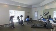 This single-floor fitness center offers a clean, functional workout environment designed to support a range of exercise routines. The space is laid out openly, allowing for easy movement between stations and equipment. Large vertical-blind-covered windows along one wall invite natural light and contribute to the room's bright, airy atmosphere. The flooring throughout is low-pile carpet in a neutral tone, providing a comfortable surface underfoot. A tray ceiling detail adds a subtle architectural element to the otherwise minimalist interior, finished in crisp white throughout. The room is equipped with a stationary recumbent bike, a rowing machine, a stair climber or elliptical-style machine, and an inversion table, offering a comprehensive range of cardiovascular and flexibility training options. A dumbbell rack with free weights and a stability ball round out the strength and core training offerings. Recessed lighting supplements the natural light, ensuring the space is well-illuminated at all times. A wall-mounted clock and an illuminated exit sign are also present. A paneled door with a window provides access to an outdoor area, further enhancing the sense of openness. Mirrored panels on one wall add depth to the room and are practical for form-checking during workouts.