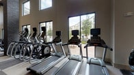 This fitness center is designed to support a full range of workout routines within a spacious, single-floor layout. The space is defined by its dramatically high ceilings, which are complemented by large floor-to-ceiling windows and elevated clerestory windows that flood the interior with natural light and offer views of the surrounding landscaped outdoor areas.

The flooring throughout consists of a distinctive checkered pattern of light and dark speckled rubber tile, providing both durability and visual interest. Dark brick columns punctuate the main workout area, adding an architectural contrast to the neutral-toned walls. Pendant light fixtures and ceiling fans contribute to both the aesthetic and functionality of the space.

The cardio zone is well-stocked with a row of Matrix treadmills and elliptical machines, each equipped with individual display screens. Strength-training options include a multi-station cable machine, a variety of Matrix selectorized equipment, and a full rack of dumbbells positioned in front of wall-length mirrors. Additional equipment includes a rowing machine, air bike, and a dedicated rack of medicine balls. Built-in shelving near the entrance provides convenient storage for personal belongings.